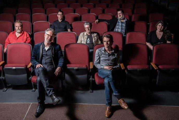 Seven artists seated in red theater rows with focus on front pair in dim cinematic setting.