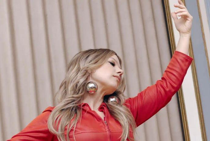 Artist in bright red outfit posing with raised arm beside gold frame and vertical beige panels.