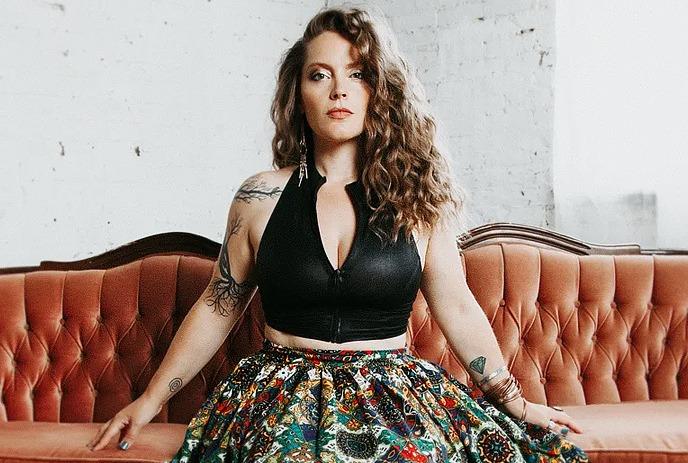 Sandra Bouza seated on a vintage rust-colored sofa, wearing a black top and patterned skirt indoors.