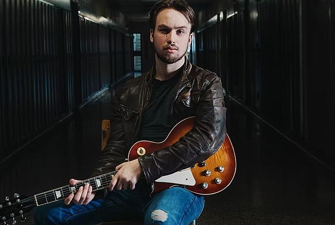 Spencer Mackenzie with a serious expression, sits in a dimly lit hallway, holding a sunburst electric guitar.