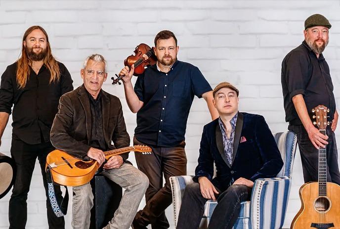 The Steel City Rovers pose against a white brick wall. They hold various instruments, conveying a relaxed, artistic vibe.