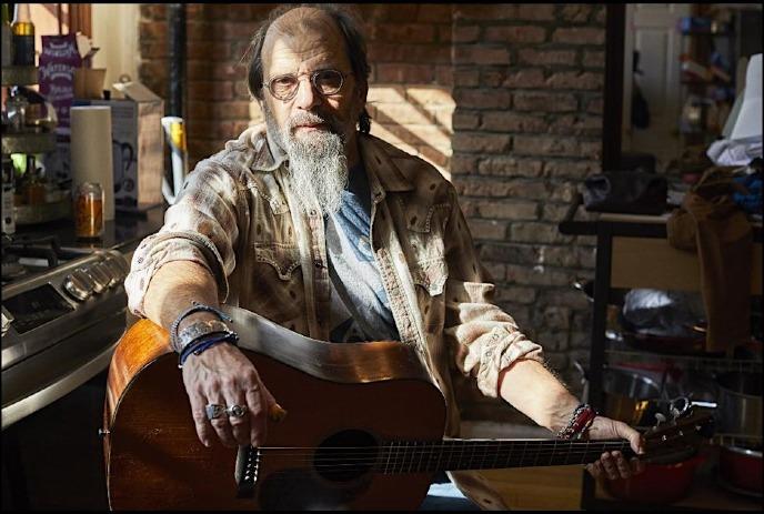 Steve Earle wearing a patterned shirt and denim jacket, adorned with multiple rings, is holding an acoustic guitar.