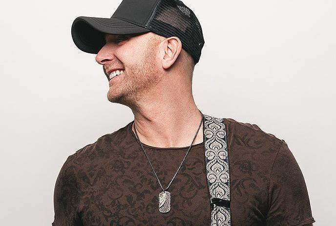 Tim Hicks posing for the camera in front of a white wall, looking to the side and holding his guitar.