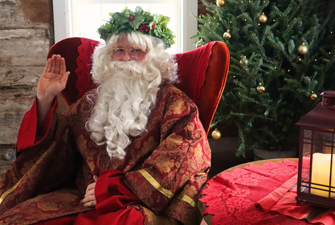 Santa figure in ornate red-gold robe and wreath, seated by candle lantern and tree, raising hand in festive gesture.