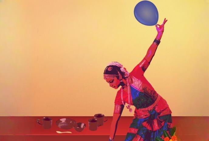 Dancer in saree holds balloon mid-pose beside tableware, set against warm gradient background.