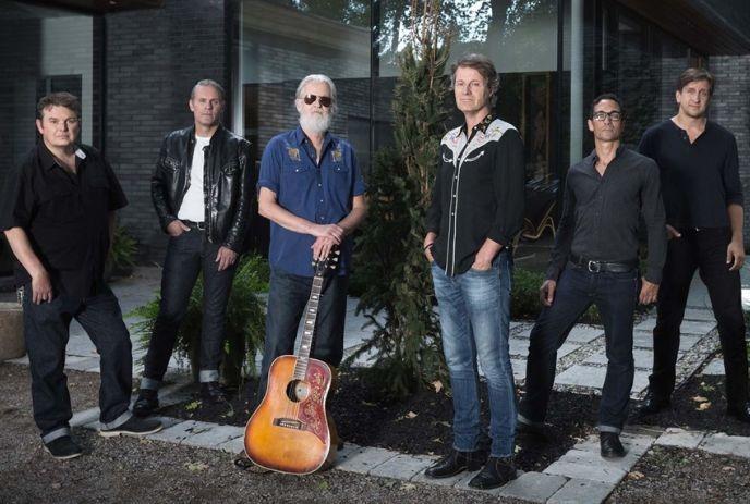 Six band members of Blue Rodeo standing outdoors in front of a modern building with large windows, posing for a picture.