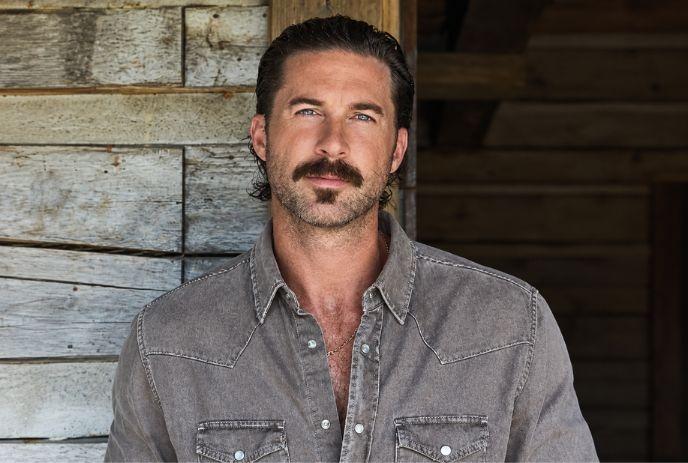 Portrait of Railey Green with a mustache wearing a gray button-up shirt, standing by a rustic wooden wall.