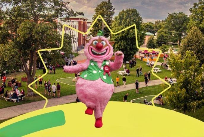 Pink mascot dances at bright outdoor fair with families tents trees and cheerful lively energy all around festival fun.