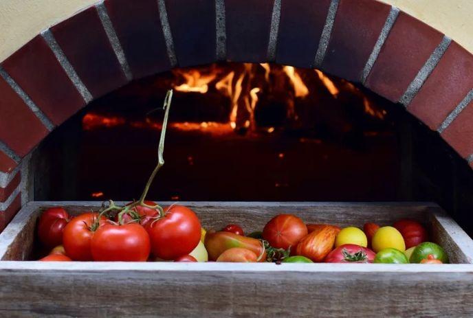 A wooden crate filled with colorful tomatoes sits in front of a brick oven with a fire burning inside.