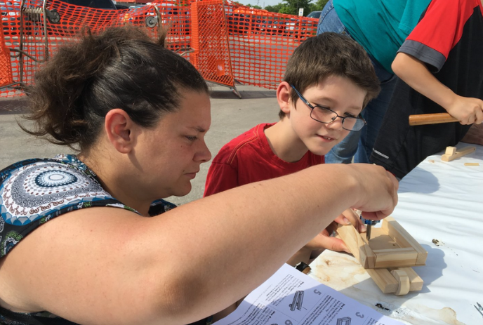 Person sitting at a table helping a child make a wooden toy, with the child looking on.