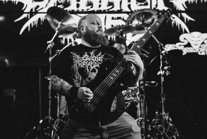 A bearded musician passionately plays a bass guitar on stage during a live metal concert performance.