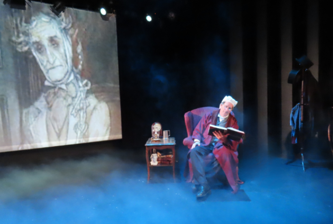 Person sitting onstage, dressed as Ebeneezer Scrooge, reading from a book.