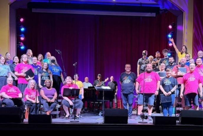 Pride chorus members stand on stage in colorful shirts, singing beneath purple stage lights.