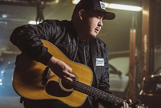 William Prince in a dark jacket, playing an acoustic guitar in a warmly lit indoor setting.