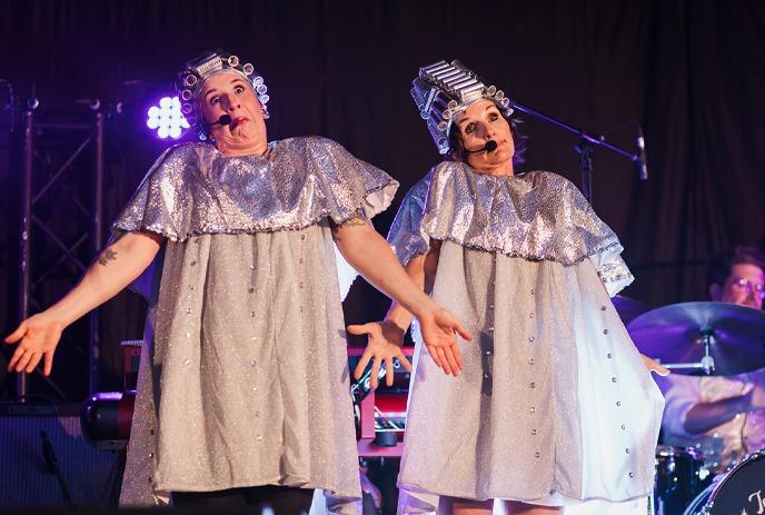 Two performers in silver outfits and hair curlers gesturing on stage.