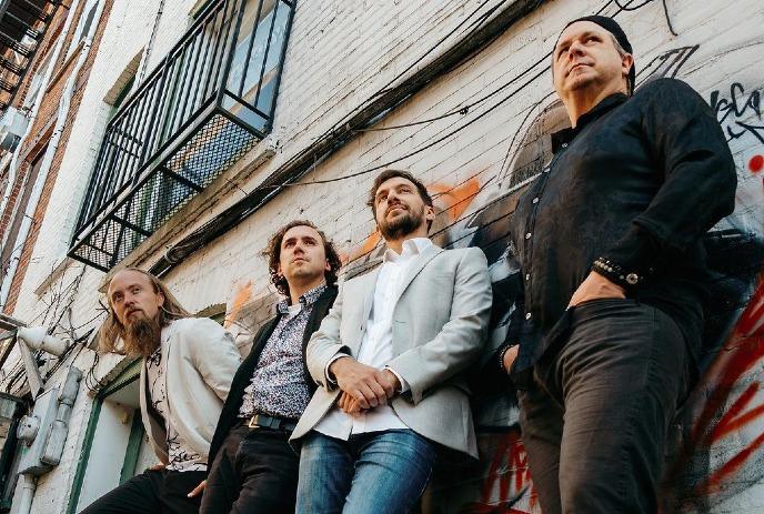 Four artists stand together with blue bass before dark backdrop featuring vivid vertical streaks of color.