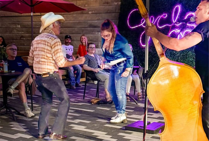 Two people dance as musician plays bass and crowd watches from nearby tables.