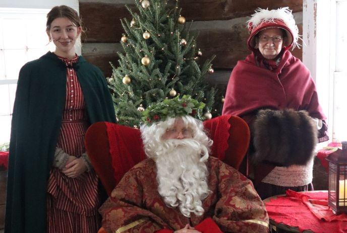 St. Nick sits between two women in 19th-century attire with a Christmas tree in the background.