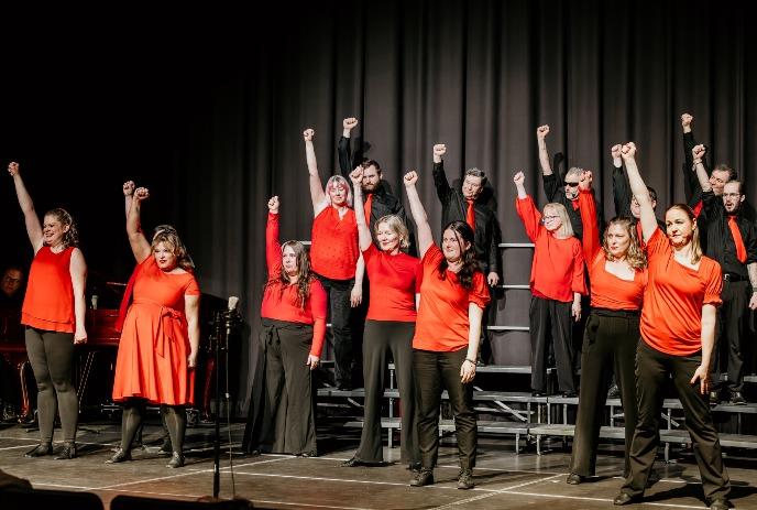 Performers in red raise fists on stage risers before dark curtain on a theatre stage.