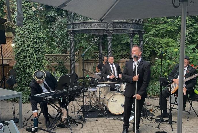 Live band in suits playing music outdoors under umbrellas, surrounded by greenery.