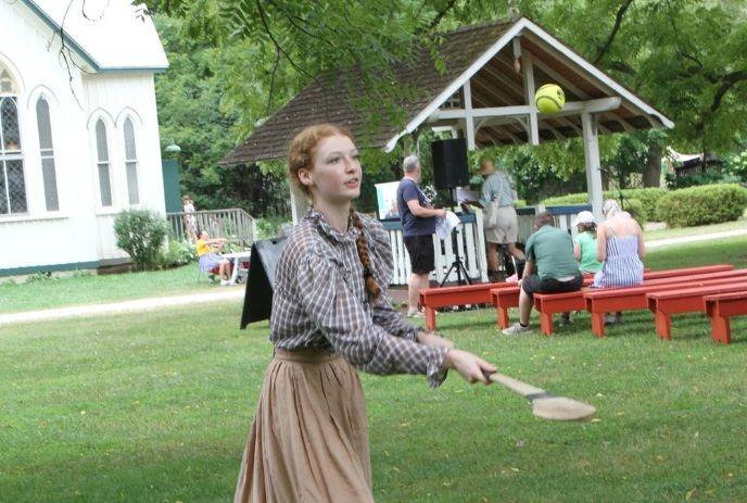 A young woman in 19th-century attire plays a lawn game at the Fanshawe Pioneer Village, with visitors gathered nearby.