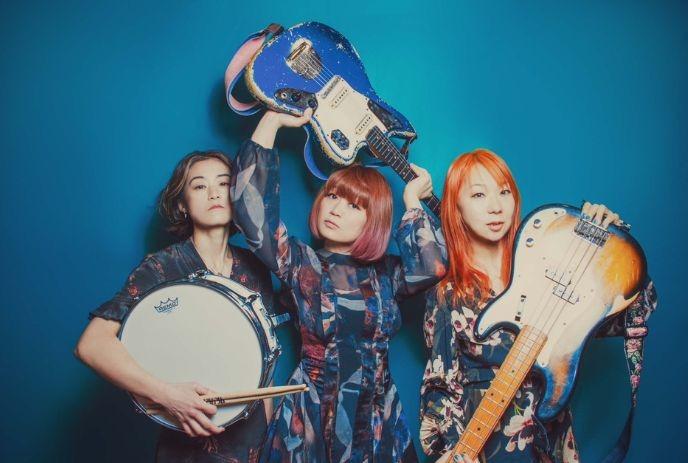 Three women artists with two guitars and one drum posing for a picture under a blue background.