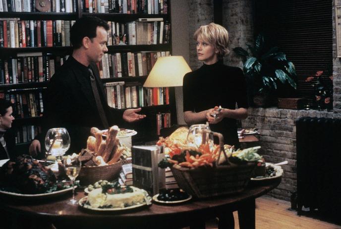 Two people standing near a table of food, bookshelves behind them, creating a cozy gathering scene.