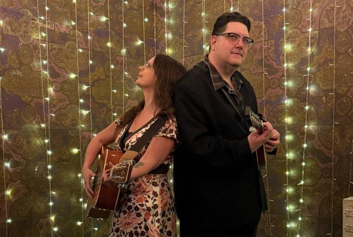 A man and a woman standing back-to-back holding guitars against string lights and ornate wallpaper.