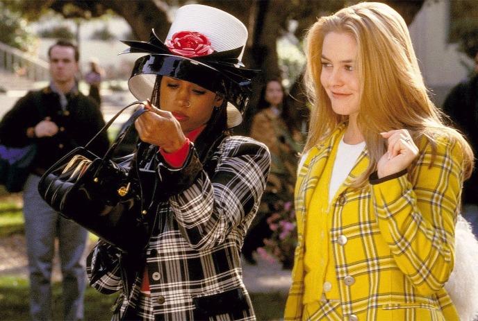 Two women in plaid coats, one yellow and one monochrome, stroll side by side on a city sidewalk.