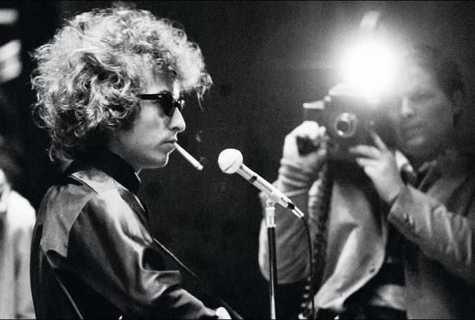 Bob Dylan in sunglasses, smoking by a microphone as a photographer snaps a black-and-white picture with a flash.