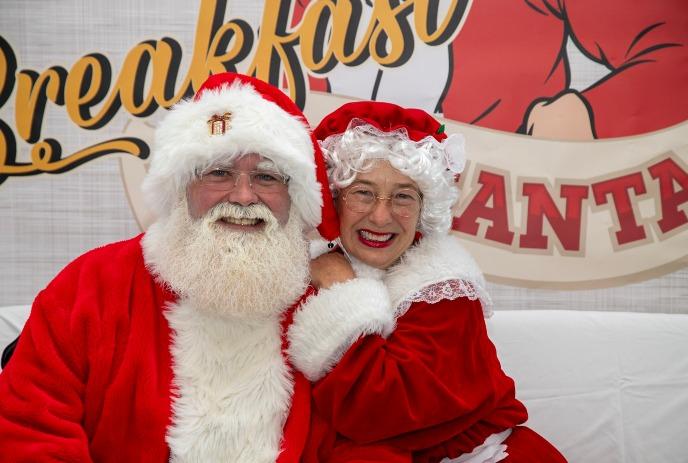 Santa and Mrs. Claus pose for a holiday photo in front of a breakfast sign.