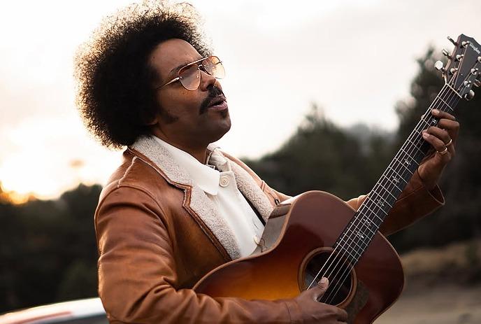 Alex Cuba playing an acoustic guitar with a sunset in the background, wearing sunglasses and a jacket.