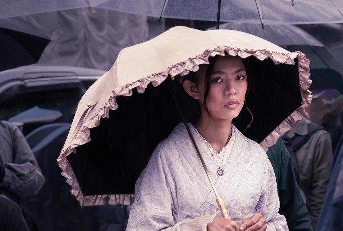 Young woman in traditional dress under a large, light pink umbrella, standing in the rain.