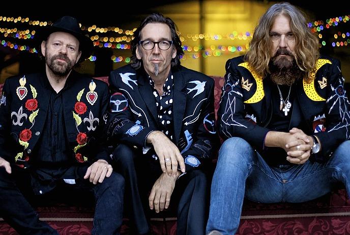 Three musicians in embroidered black jackets and sunglasses pose confidently against a bold, colorful geometric backdrop.