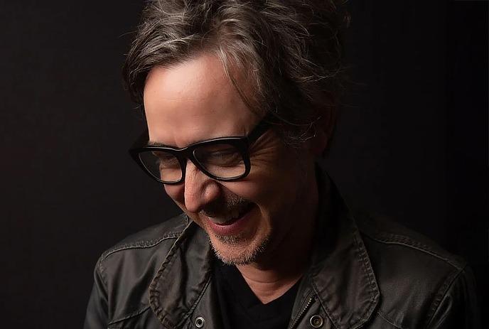 A black and white photo of Danny Michel smiling with his head turned from the camera, wearing a hat against a white background.
