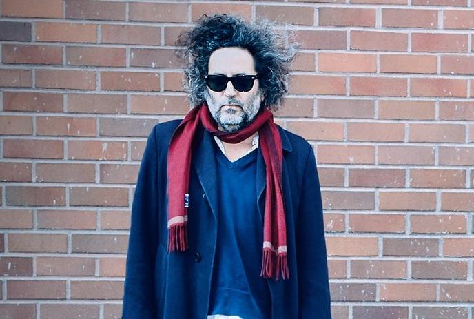 Destroyer leaning on stone wall in a rustic setting, wearing a blazer and t-shirt.