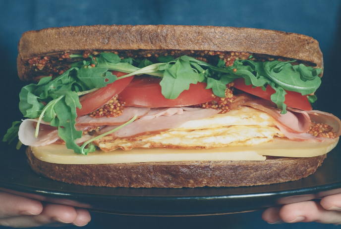 Close-up of a sandwich on whole grain bread with cheese, fried egg, ham, tomato, arugula, and mustard.