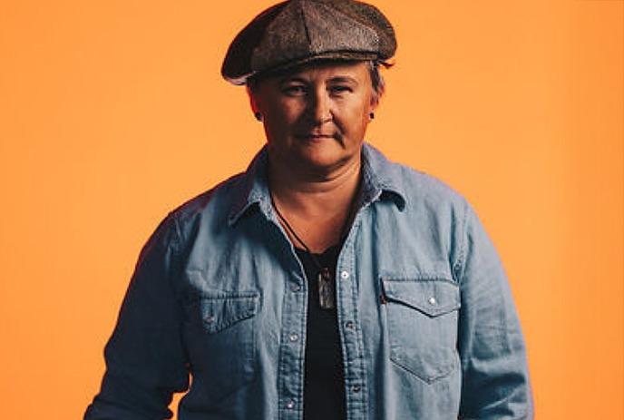 Irish Mythen stands confidently in a denim shirt and cap against an orange backdrop