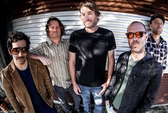 The band Mad Caddies stands in front of a corrugated metal wall, dressed in casual attire.