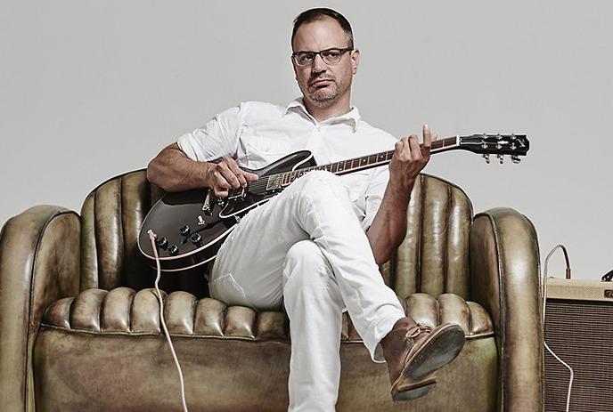 Musician Matthew Good sitting on a couch holding an electric guitar plugged into an amp.