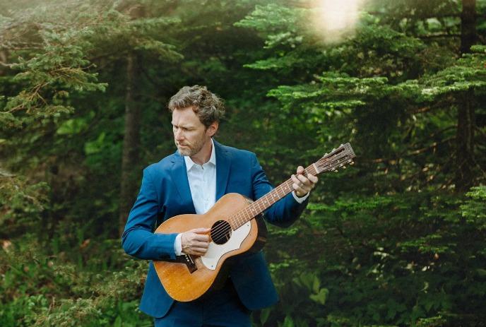 Ian Sherwood holding an accoustic guitar, with green pine trees in the background.