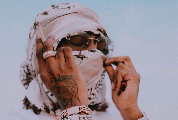 NORTHSIDEBENJI wearing a white headscarf and jewelry, holds a cigarette.