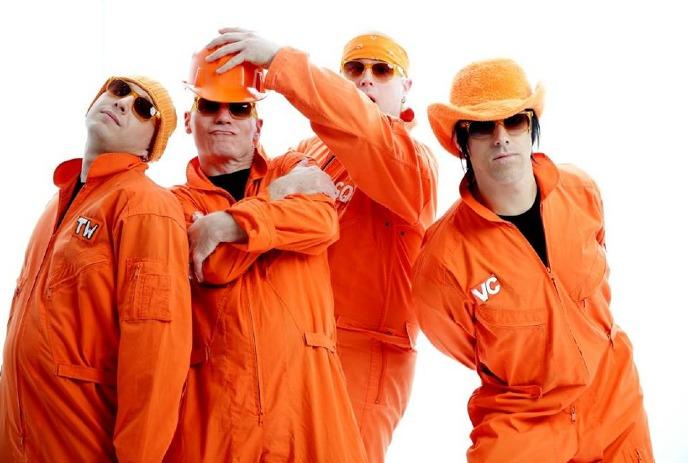 Four persons posing for a photo against a white background while wearing orange clothing, headgear, and glasses.