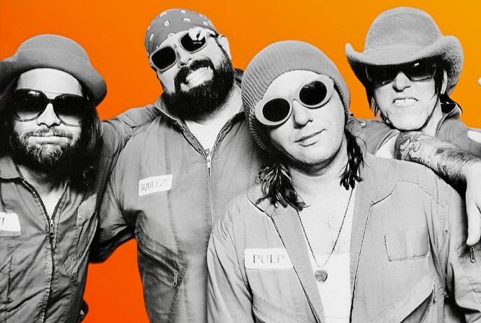 Four band members in matching jumpsuits pose before a bold orange circle, posing for a picture.