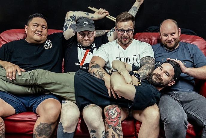 Five friends on red couch with one lying across laps as others pose playfully against dark backdrop.