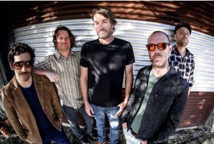 The band Mad Caddies stands in front of a corrugated metal wall, dressed in casual attire.
