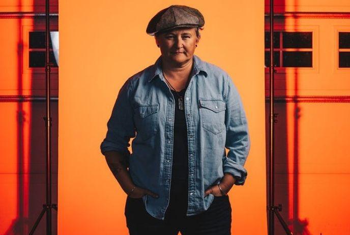 Irish Mythen stands confidently in a denim shirt and cap against an orange backdrop