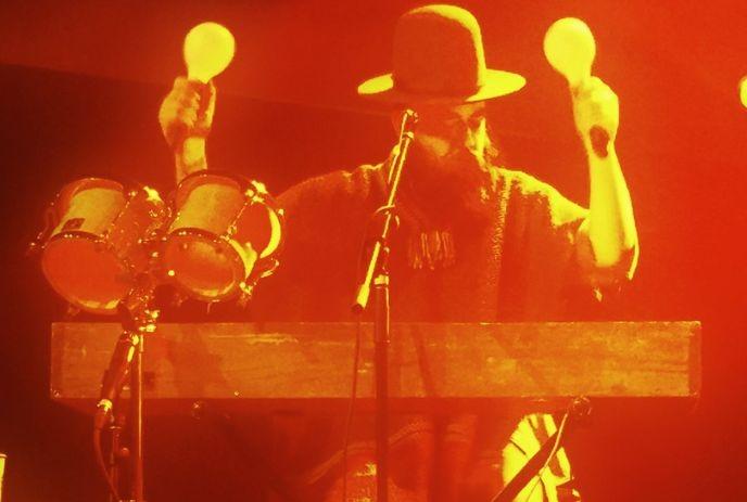 A musician in a hat and poncho plays maracas on a red-lit stage, standing behind drums.