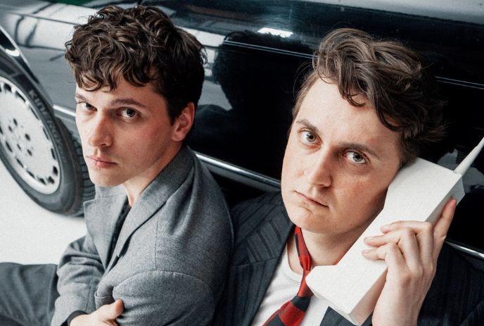 Two people in suits sit near a car. One holds a vintage mobile phone, posing for the picture.