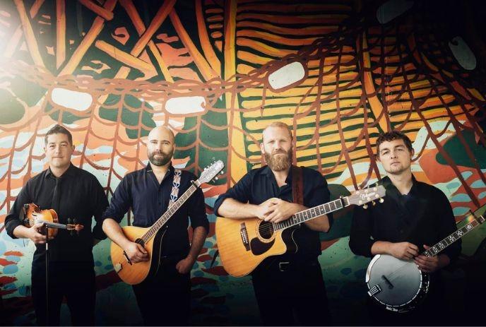 Rum Ragged band standing together, each holding an instrument, posing in front of a vibrant, colourful wall.